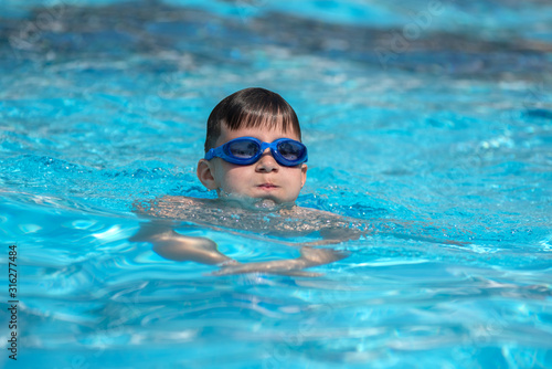 European boy in swimming goggles is swimming in the hotel’s pool during his summer vacations.