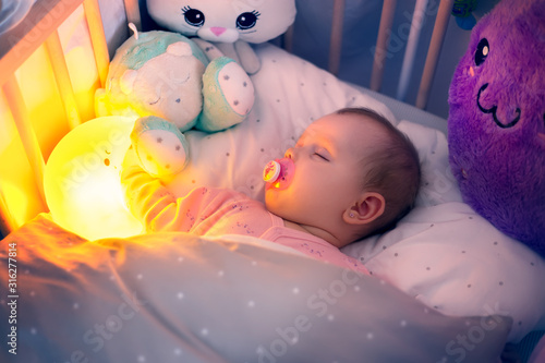 Baby sleeps in bed with lighting moon and soft toys in the night