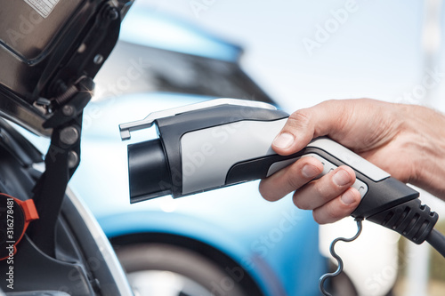 Transportation. Young man traveling by electric car stop at chraging station holding charging plug close-up