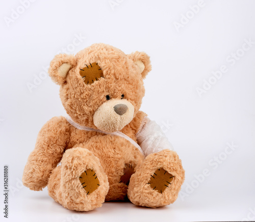 large beige teddy bear with patches sits on a white background, left paw is bandaged with a white medical bandage