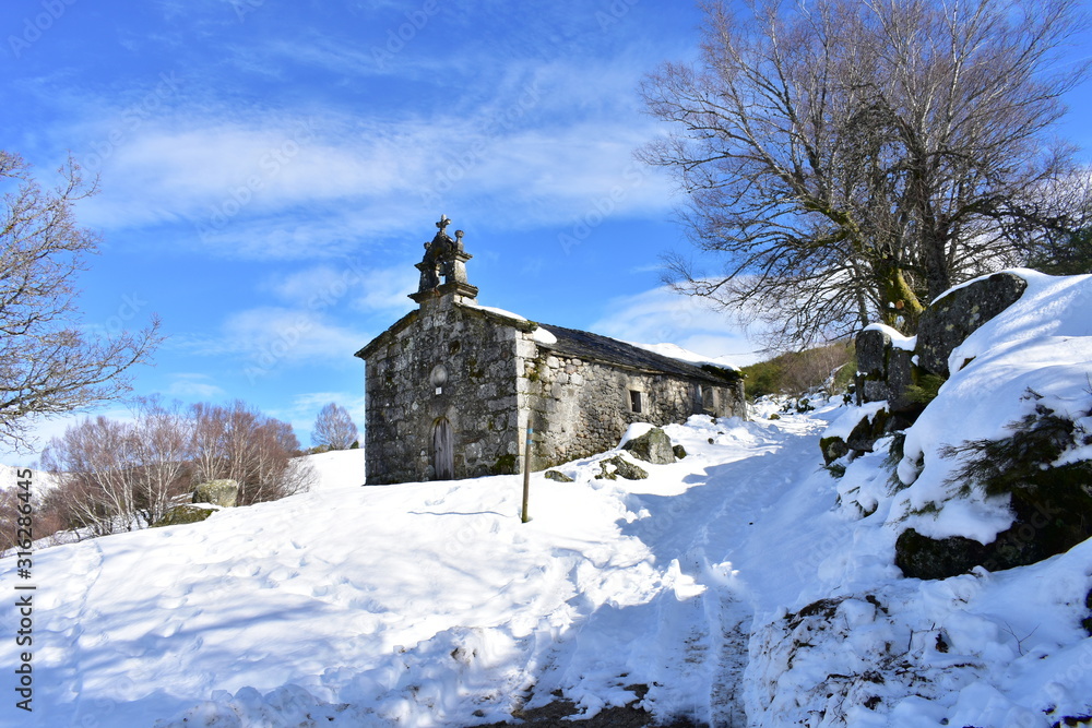 Ermita de San Lorenzo with snowy slope, trees and blue sky. Piornedo Village, Ancares, Lugo, Galicia, Spain.
