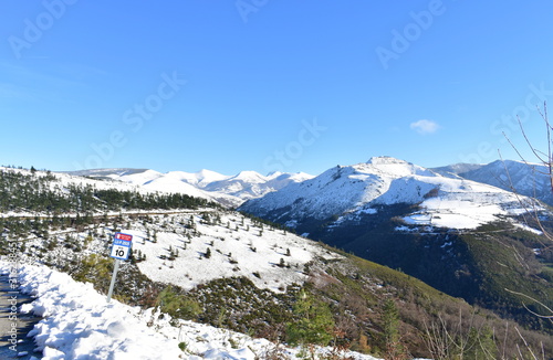 Winter landscape with snowy mountain range and green valley with forest. View from a mountain road with blue sky. Ancares Region, Lugo Province, Galicia, Spain.