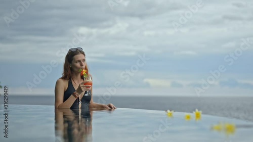 Beautiful young red haired woman drinking exotic cocktail in ocean pool at bali resort. Female enjoying beverage and watching amazing natural scenery at poolside under blue sky. Brunette wearing