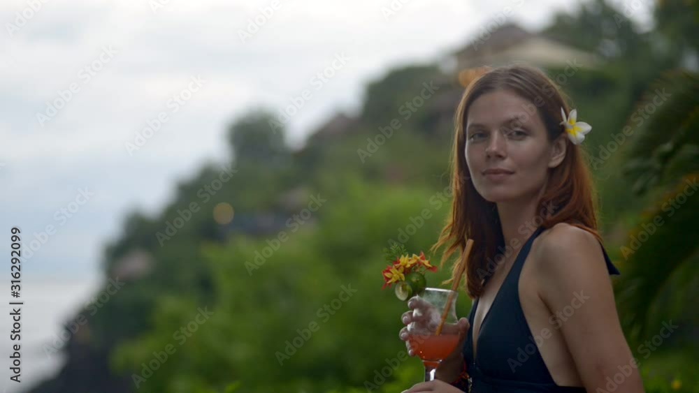 Beautiful young red haired woman posing with cocktail in hand while standing on tropical island. American female model looking at camera with happy smile having time at luxury resort. Portrait of lady Beautiful young red haired woman posing with cocktail in hand while standing on tropical island. American female model looking at camera with happy smile having time at luxury resort. Portrait of lady