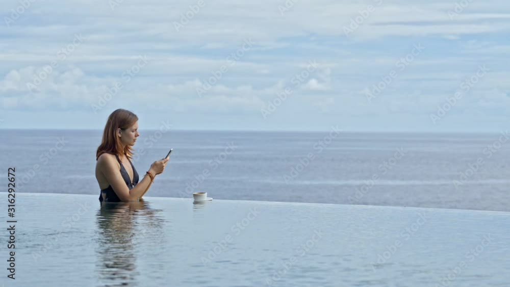 woman in infinity pool with coffee and holding mobile phone to take picture. pretty sexy female relax on sea ocean horizon. take photo panorama on vacation in bali hotel exotic resort woman in infinity pool with coffee and holding mobile phone to take picture. pretty sexy female relax on sea ocean horizon. take photo panorama on vacation in bali hotel exotic resort