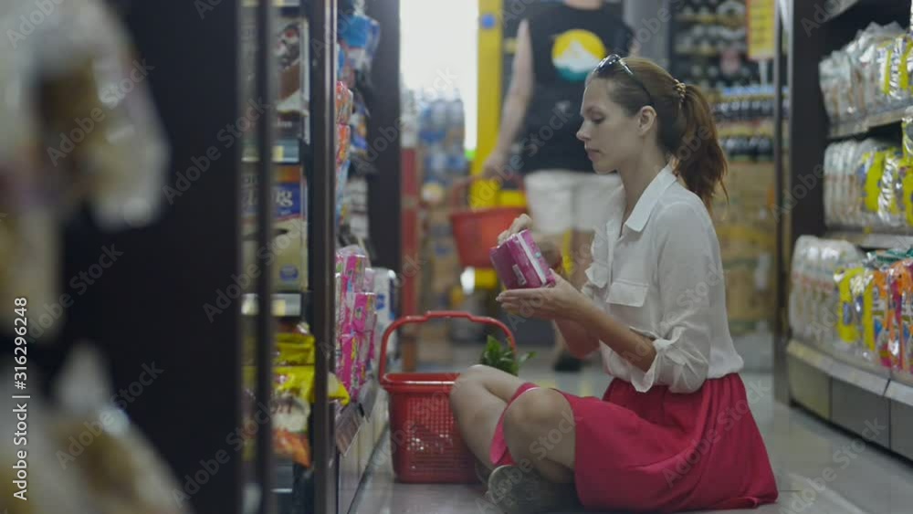 Young american woman is choosing female hygiene goods pads sitting on supermarket floor. Side of brunette customer holding package in hand, reading and putting in basket. Young american woman is choosing female hygiene goods pads sitting on supermarket floor. Side of brunette customer holding package in hand, reading and putting in basket.