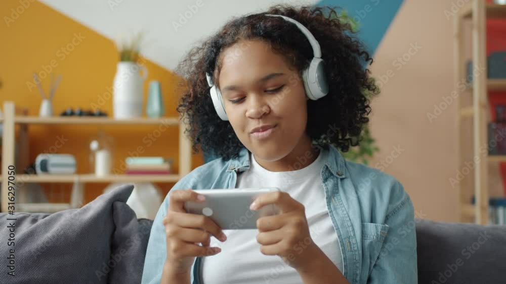Afro-American teenager is playing smartphone game and listening to music at home wearing wireless headphones. Entertainment and modern devices concept. Afro-American teenager is playing smartphone game and listening to music at home wearing wireless headphones. Entertainment and modern devices concept.