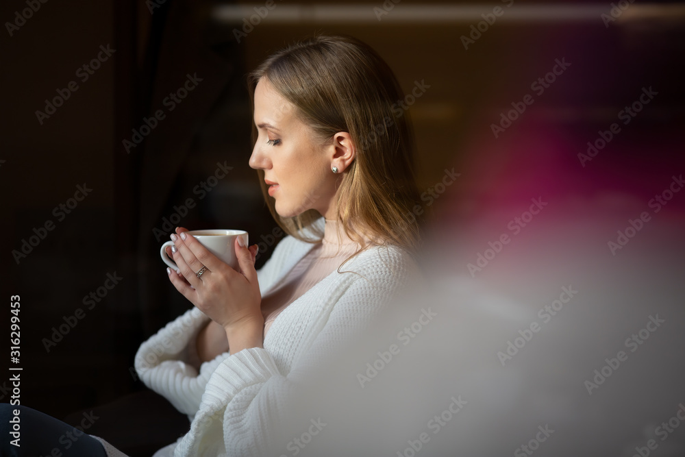 Beautiful girl with brown hair is drinking coffee. Attractive young woman in a white sweater with a cup of coffee in her hands is relaxing in a cozy cafe.