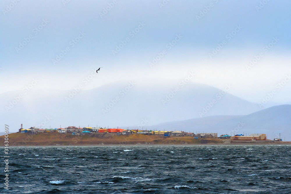 View from the sea to the Chukchi village of Yanrakynnot, located on the shores of the Senyavin Strait of the Bering Sea, surrounded by tundra and mountains. Autumn rainy weather. Chukotka, Russia. View from the sea to the Chukchi village of Yanrakynnot, located on the shores of the Senyavin Strait of the Bering Sea, surrounded by tundra and mountains. Autumn rainy weather. Chukotka, Russia.
