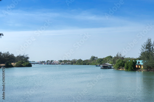 Houseboat in the river, near the shore, with green trees, behind a small fishing boat is parked. And far away are many fishing boats and a fishing community near the sea