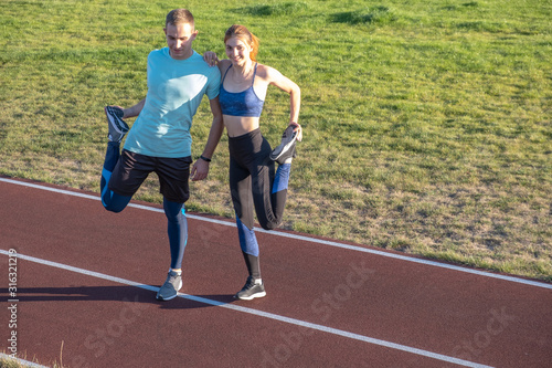 Young couple of fit sportsmen boy and girl doing warming up exercises  before running on red tracks of public stadium outdoors. © bilanol