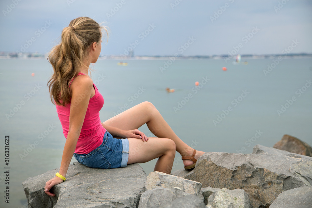 Young woman in summer clothes sitting on big boulders on sea shore looking on horizon.