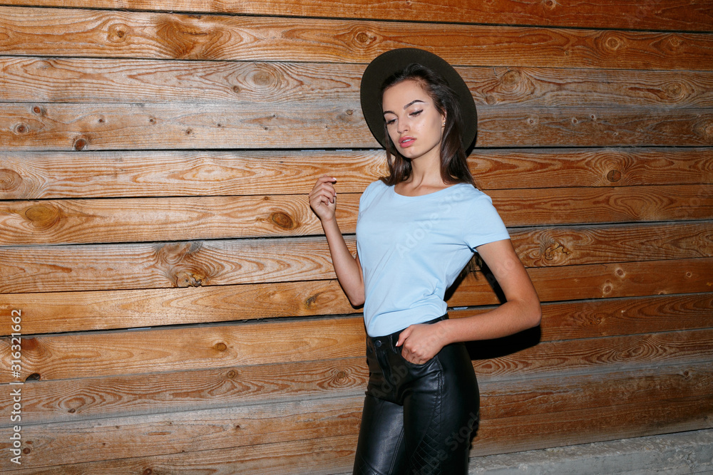 Attractive girl in hat, with light on it, wear in stylish clothes, posing near wooden wall, looking seductive at camera, with hands in her pocket. Copy space. Attractive girl in hat, with light on it, wear in stylish clothes, posing near wooden wall, looking seductive at camera, with hands in her pocket. Copy space.