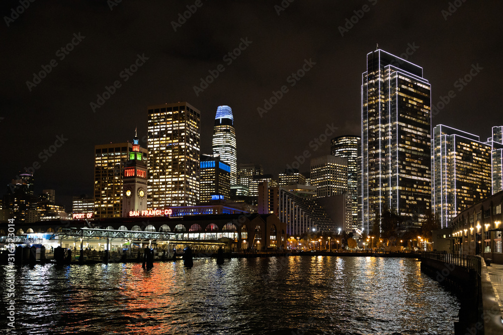San Francisco port illuminated at night, against the backdrop of impressive skyscrapers with light and reflection in the water, view from the side of the San Francisco Bay.