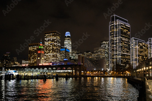 San Francisco port illuminated at night, against the backdrop of impressive skyscrapers with light and reflection in the water, view from the side of the San Francisco Bay. © Volodymyr