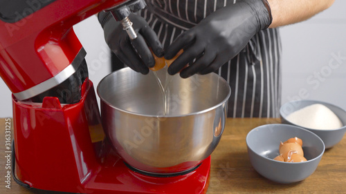 A male pastry chef breaks chicken eggs. The red kitchen mixer is designed to produce uniform consistency.