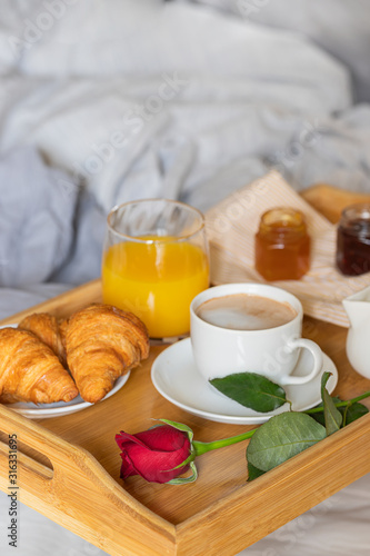 Breakfast on the bed inside a bedroom. Wooden try with coffee, orange juice, croissant with jam and red rose flower. Romantic surprise.