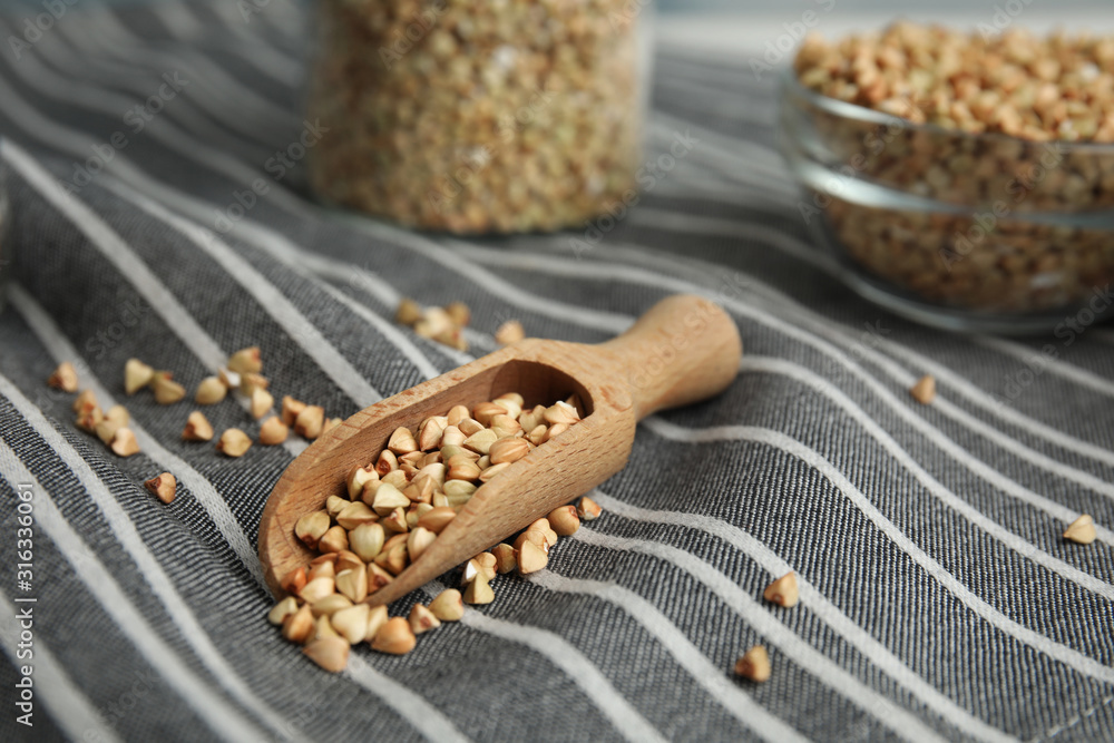 Fototapeta premium Uncooked green buckwheat grains in scoop on fabric, closeup