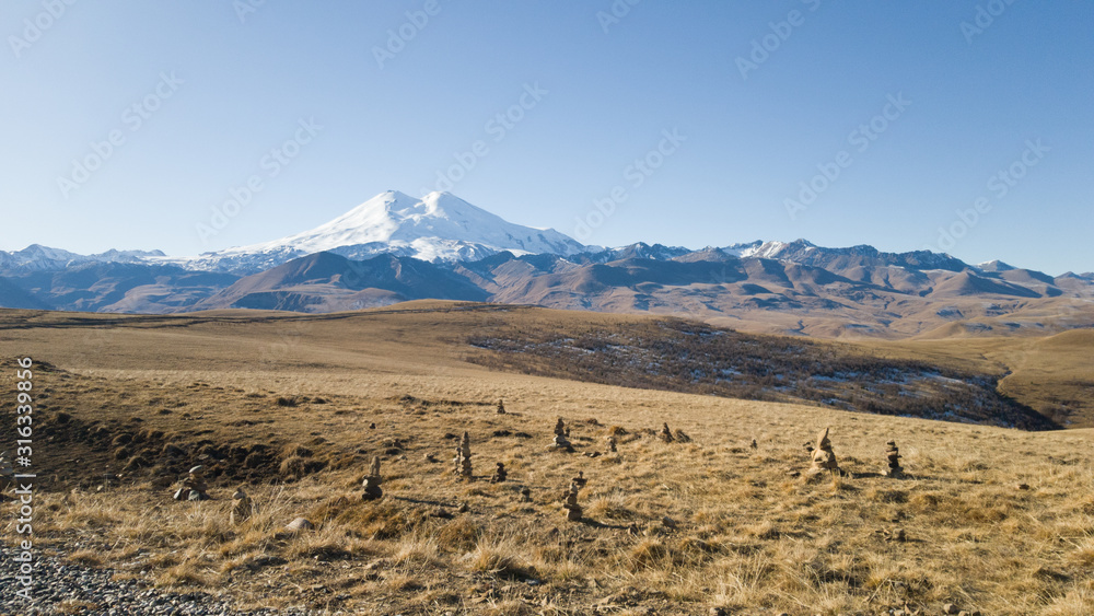 Autumn landscape. View of the dry yellow field and the beautiful mount Elbrus in the Caucasus. Pyramids of stones in the foreground. Travel to Russia