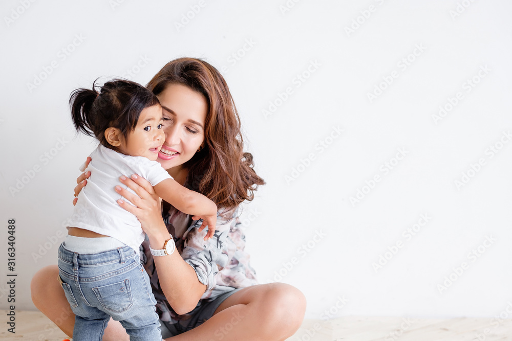 Young happy mother holds her charming mixed-race daughter and poses on a white wall background. Concept of a loving and caring family. Advertising space Young happy mother holds her charming mixed-race daughter and poses on a white wall background. Concept of a loving and caring family. Advertising space