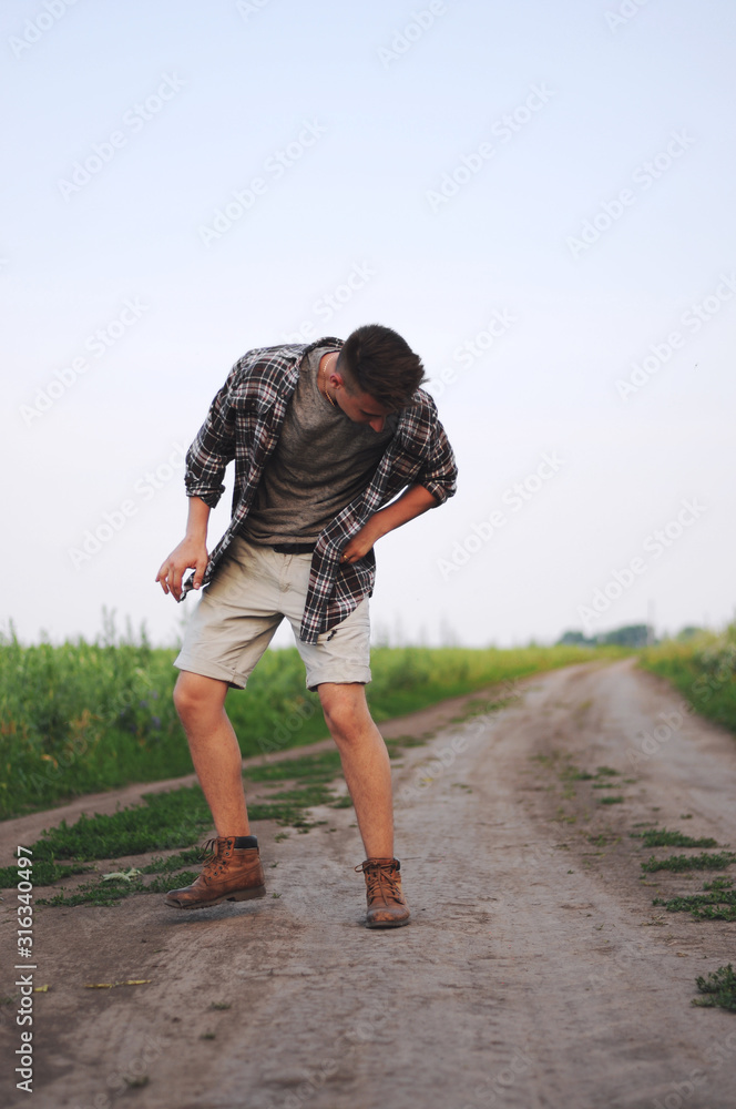 A man in a plaid shirt stands along a dirty road in the countryside.Conceptual photo of traveler.