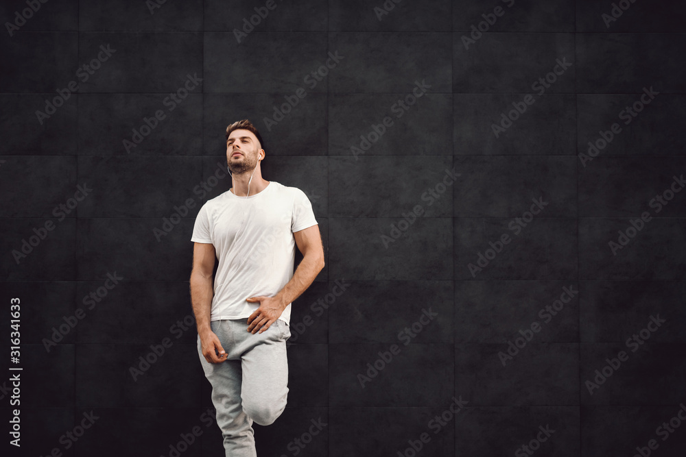 Attractive fit caucasian sportsman in tracksuit and in t-shirt leaning on the wall and resting. In ears are earphones.