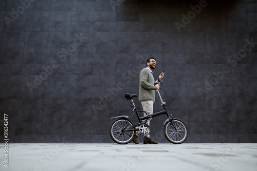 Full length of smiling caucasian fashionable man pushing bicycle and holding smart phone while passing by gray wall.