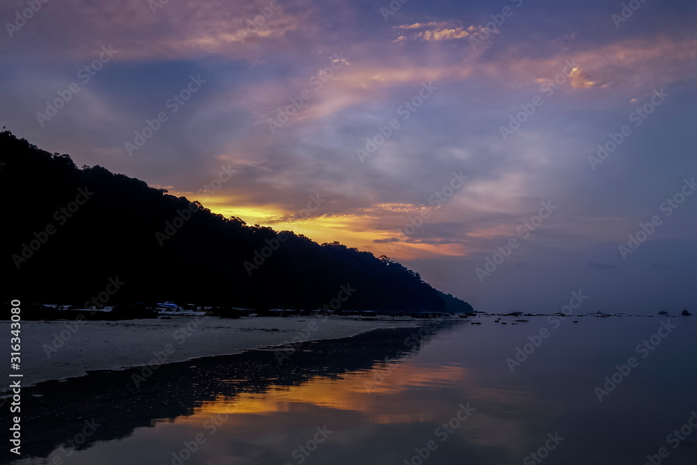 Sea view morning of white sand beach with mountain around with soft mist and orange sun light in cloudy sky background, twilight at Khao Chong Kad, Surin island, Mu Ko Surin National Park, Thailand.