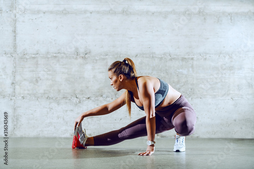 Fit attractive caucasian brunette in sportswear with ponytail crouching and stretching leg. In background is wall.