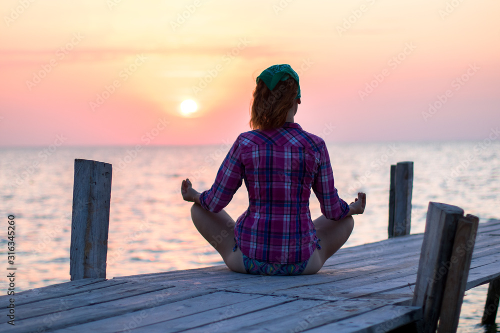 girl is sitting in lotus pose on an old wooden pier and enjoys the sunset on the sea