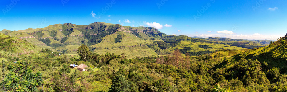 Panoramic view over a forest with a little hut embedded and green mountains, sunny day, Drakensberg, Giants Castle Game Reserve, South Africa