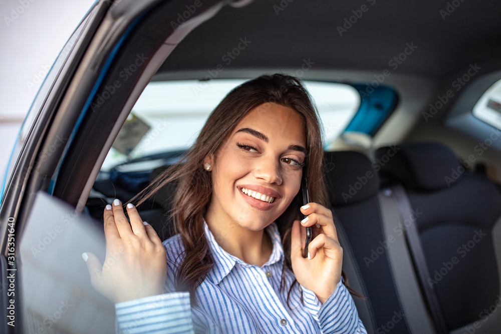 Young businesswoman talking on the phone in the back seat of the car. Young smiling woman holding smartphone in her hand and talking on the back seat in car.