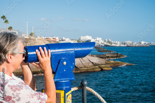 Side view of a senior woman with gray hair looking at the sea with a touristic binocular. Blue sky and ocean © luciano