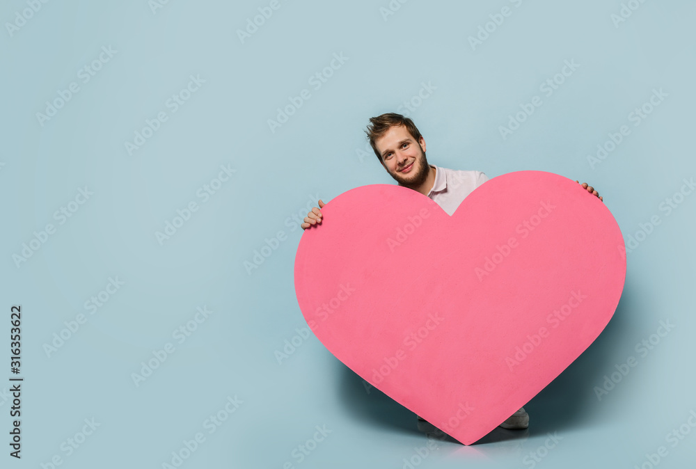 Love gift for valentines day. Young man standing with pink heart shape toy with hands on pastel blue Love gift for valentines day. Young man standing with pink heart shape toy with hands on pastel blue