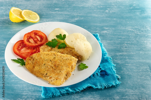 Close-up of a healthy baked battered fish, seasoned with fresh parsley and lemon and accompanied by cooked potatoes and tomato. Blue background. Concept of nutritious food for a healthy diet.