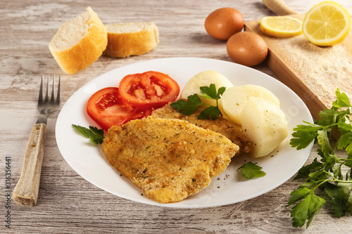 Closeup of preparation of a healthy fish with egg and breadcrumbs cooked in the oven. It is seasoned with fresh parsley and lemon and accompanied by cooked potatoes and tomatoes.