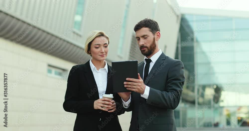 Portrait shot of the young Caucasian handsome businessman showing something to his female colleague on the tablet computer and explaining her something. Outdoor. Portrait shot of the young Caucasian handsome businessman showing something to his female colleague on the tablet computer and explaining her something. Outdoor.