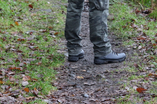 A photograph of a man's legs, standing on a forest woodland path, wearing camouflage trousers and black hiking boots. Outdoors, hiking, nature concept.
