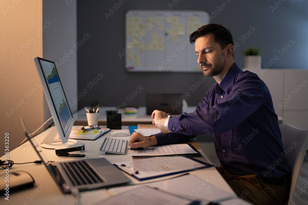 Businessman Looking At Watch While Working Late Businessman Looking At Watch While Working Late