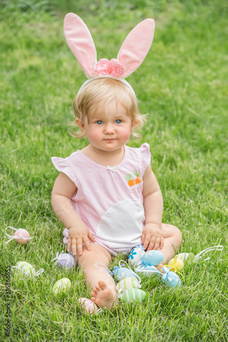 Adorable blonde toddler girl wearing bunny ears playing with Easter eggs in a basket sitting in a sunny garden