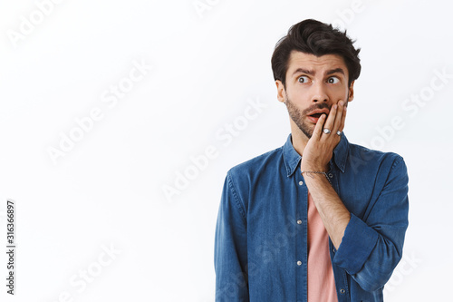 Concerned, worried young caucasian man with beard, touch cheek and look away alarmed, nervously thinking about something bad happened, gasping standing uneasy, pondering what to do with problem