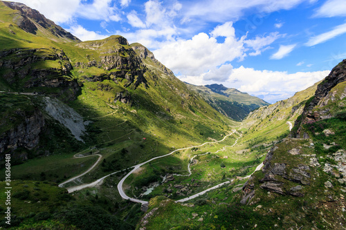Foto Panoramic view of a valley in the Pennine Alps with a winding road, green alpine pastures under a blue sky with some white clouds on a summer day