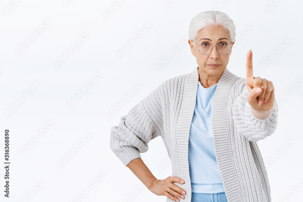 Stop right there young man. Serious-looking strict and displeased grandmother, old lady with grey hair and glasses, raise one finger in warning or prohibition gesture, scolding someone bad behaviour Stop right there young man. Serious-looking strict and displeased grandmother, old lady with grey hair and glasses, raise one finger in warning or prohibition gesture, scolding someone bad behaviour