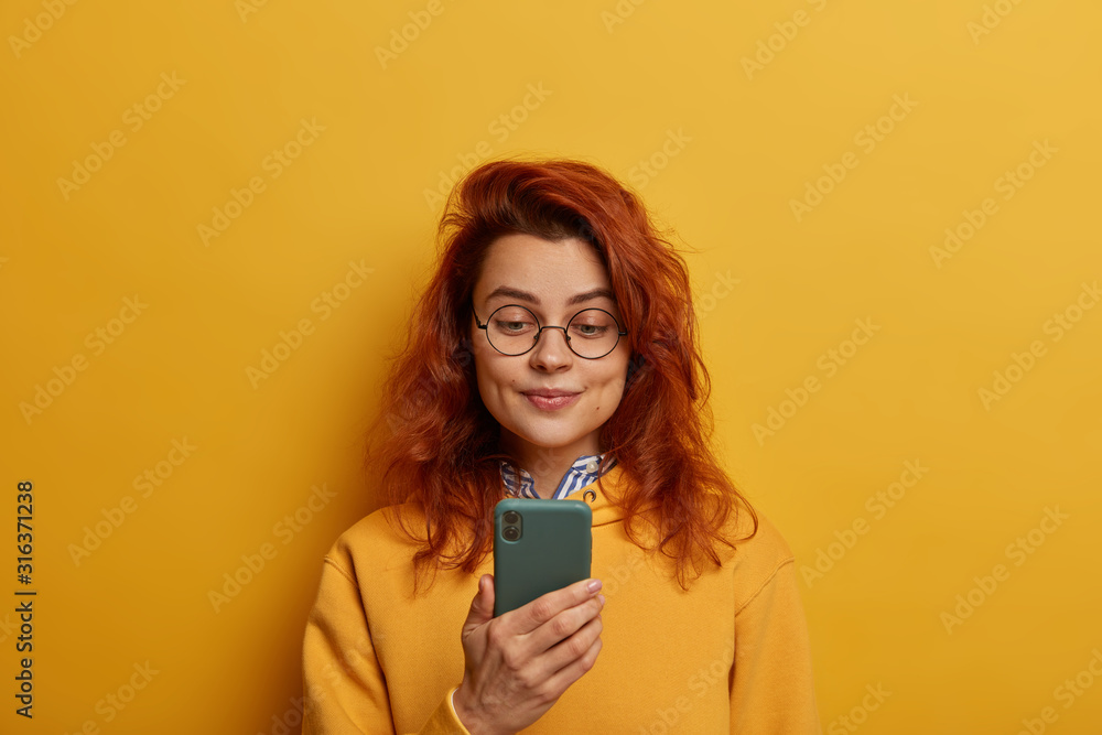 Ginger young woman holds mobile phone, reads notification, wears round glasses and yellow jumper, connected to wireless internet, isolated over yellow wall. Monochrome. People and technology concept