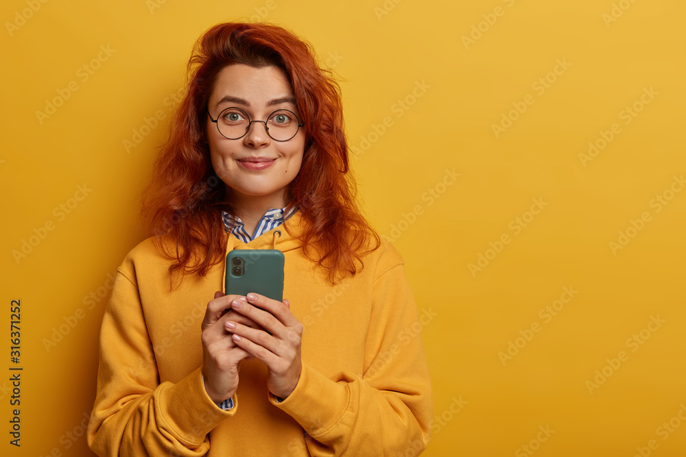 Lovely young woman has ginger hair, holds mobile phone for sending messages and surfing social networks, wears sweatshirt, round glasses, stands against yellow vibrant background, empty space Lovely young woman has ginger hair, holds mobile phone for sending messages and surfing social networks, wears sweatshirt, round glasses, stands against yellow vibrant background, empty space