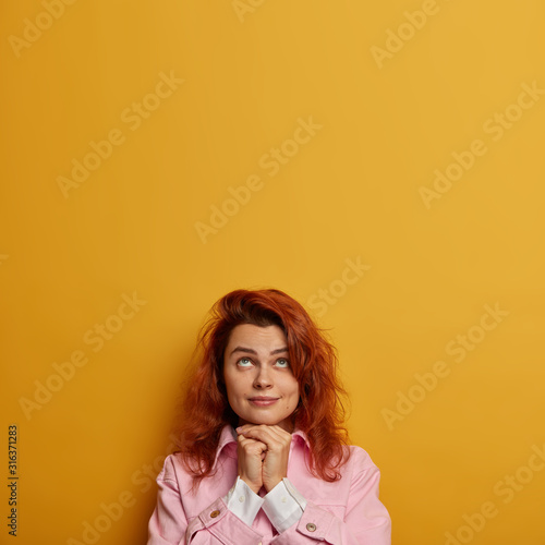 Vertical image of pensive lovely woman looks hopefully above, keeps hands under chin and prays for good wellness, has red hair, wears fashionable clothes, stands over vivid yellow background © wayhome.studio 