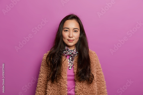 Serious dark haired woman in brown coat, has healthy skin, minimal makeup, looks with charming expression, has straight gaze at camera, isolated over lilac background. Asian female in outerwear