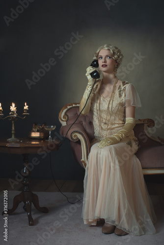 Retro 1920s fashion woman on the phone while sitting on sofa beside table with candlestick and glass of champagne.