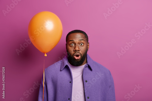 Indoor shot of speechless bearded Afro American guy opens mouth widely, cannot believe own eyes, hears bad rumors, forgets to congratulate friend in time, poses with balloon against vivid background