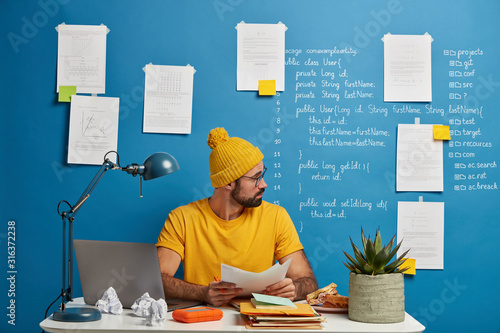 Inexperienced European man looks through paper documents, analyzes during working process, wears yellow hat and t shirt, sits at desktop against blue background with papers and written information photo